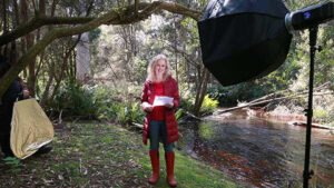 Rini in a red coat and boots standing on a river bank with a film crew